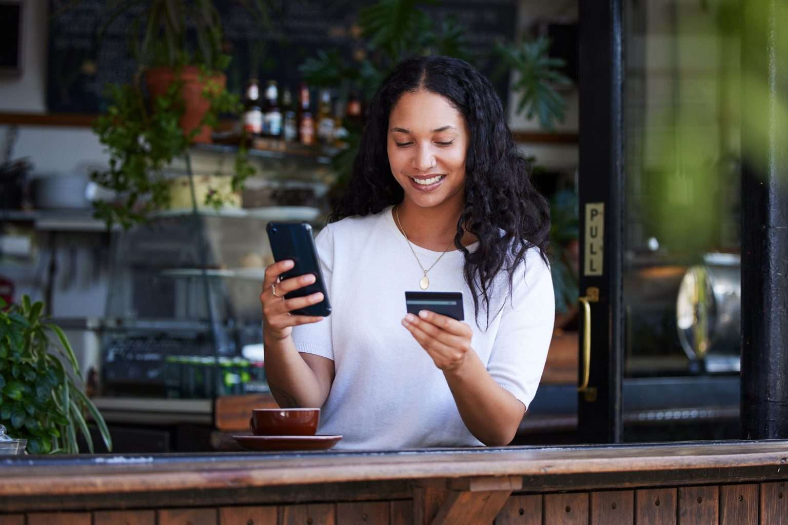 Smiling woman shopping online with her phone and credit card at a café counter