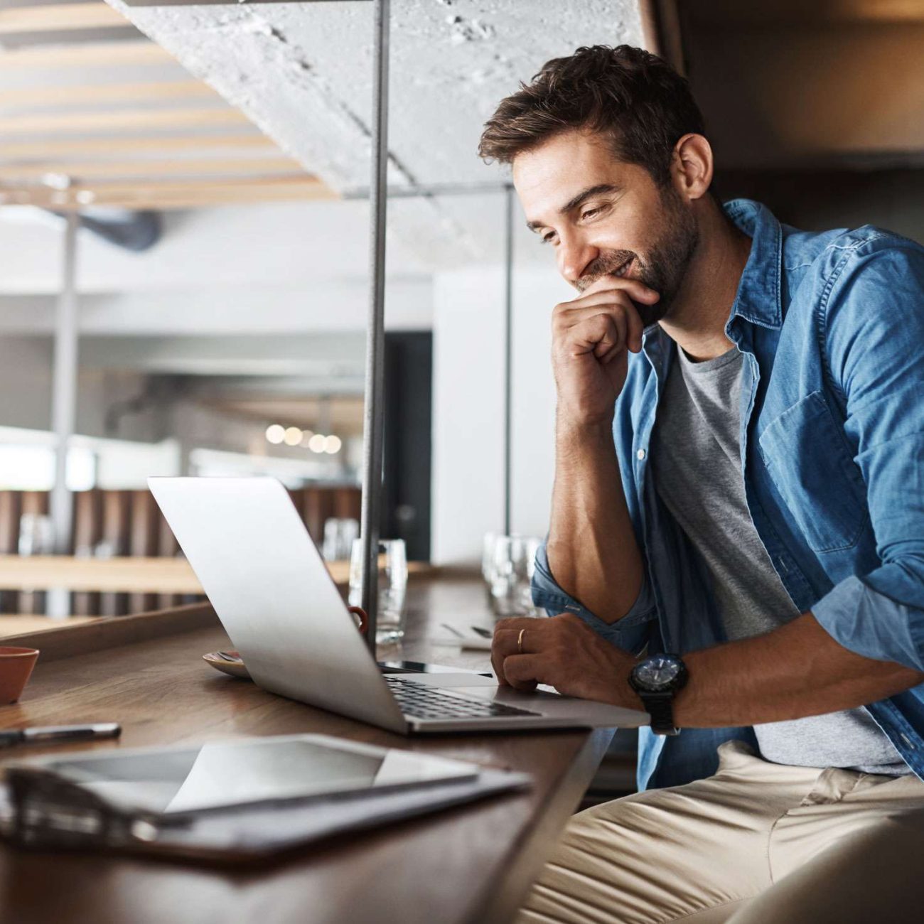 Smiling man in a denim shirt working on his laptop in a bright café.