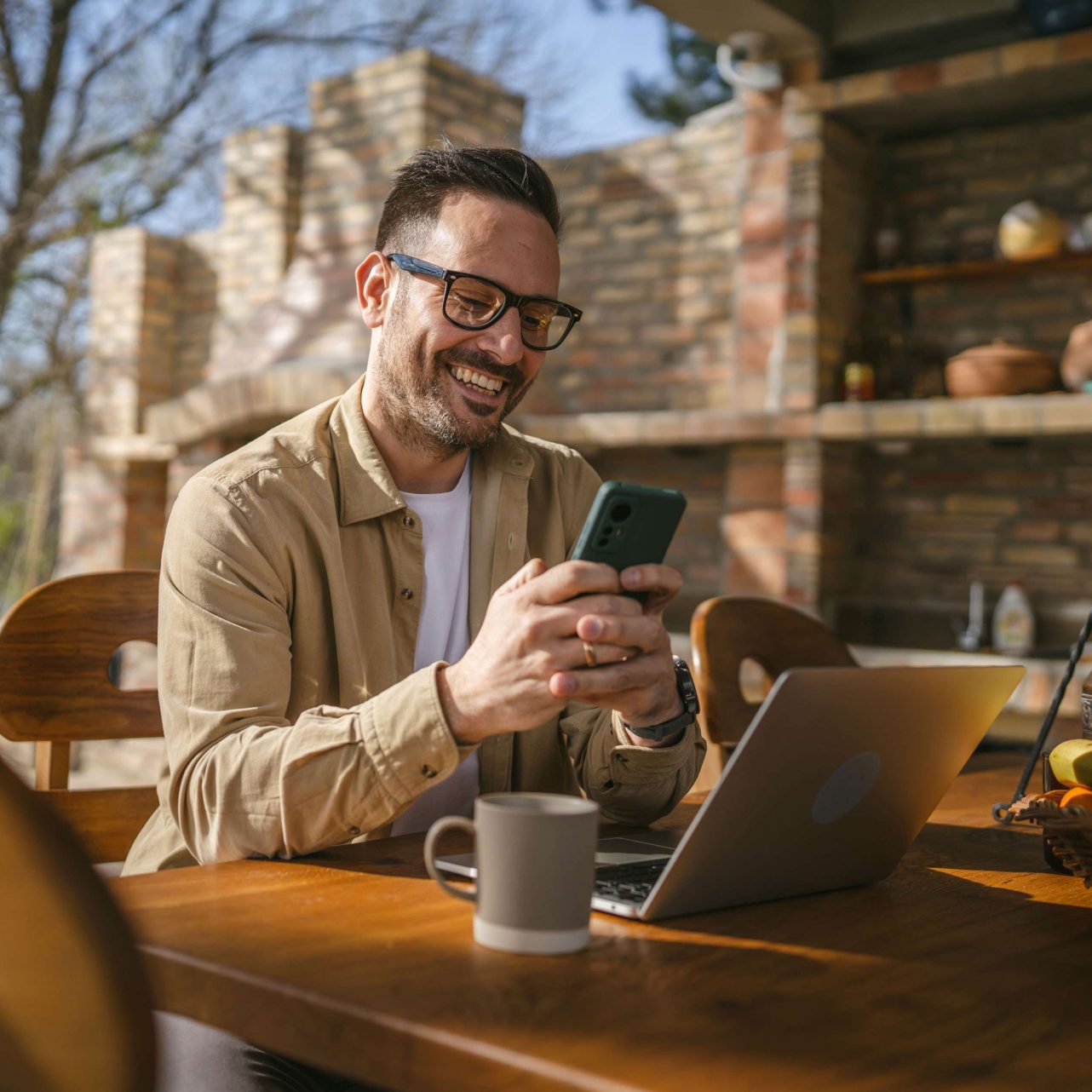 Smiling small business owner checking his phone at an outdoor table with a laptop and coffee